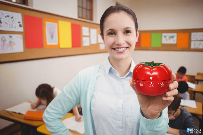 Professora sorridente em sala de aula segura um cronômetro Pomodoro vermelho ajustado para 25 minutos, enquanto alunos ao fundo realizam atividades escolares.